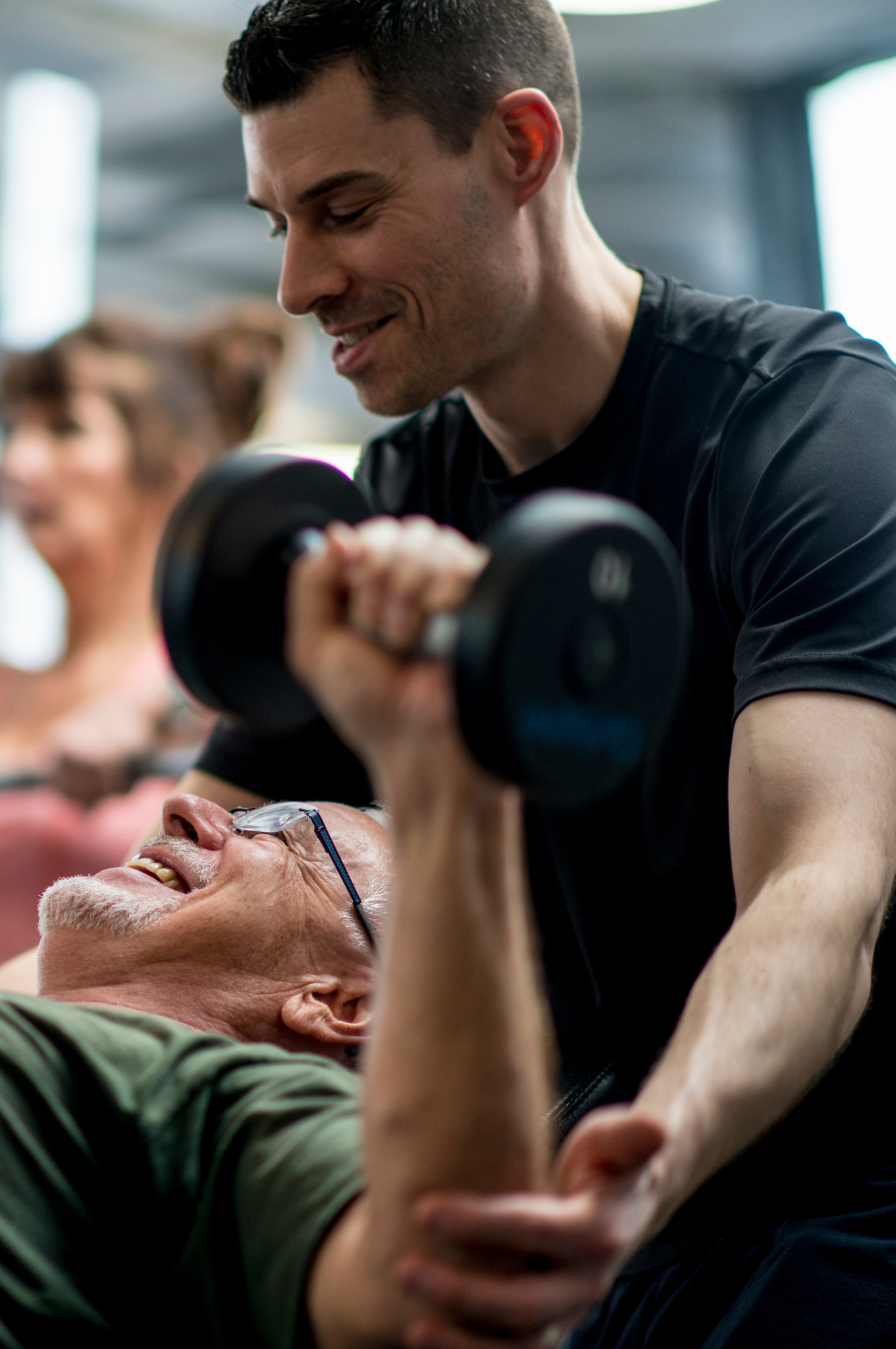 Personal trainer auxiliando idoso em treino de musculação, destacando a importância da Educação Física na qualidade de vida na terceira idade.
