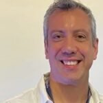 Dr. Luiz Canevali, homem de pele clara e cabelos grisalhos curtos, sorri amplamente para a câmera com os dentes à mostra. Ele veste camisa social branca com os primeiros botões abertos e usa um crachá azul pendurado no pescoço com a identificação da Faculdade Phorte. A imagem está centrada em seu rosto e parte superior do corpo, com fundo branco liso e iluminação clara. Sua expressão transmite simpatia e acolhimento, em um ambiente típico de evento educacional.