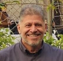 Professor Mario Charro, homem de pele clara, cabelos curtos e grisalhos e barba por fazer, sorri amplamente com os dentes à mostra. Ele veste camisa social cinza escura abotoada até o pescoço. Ao fundo, há uma placa preta de sinalização com ícones brancos indicando elevador e acessibilidade, sugerindo ambiente interno em área de circulação. O professor está parcialmente abraçado por alguém fora do enquadramento, em clima descontraído.
