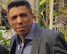 Dr. Milton Rocha, homem de pele parda, cabelos curtos e escuros, sorri levemente enquanto posa para a foto. Ele veste uma camisa azul social com botão aberto na gola, blazer cinza escuro e jeans. No pescoço, usa um crachá preto com identificação do “20º Encontro Internacional Phorte”. Ele está ligeiramente inclinado para o lado, com o braço apoiado nas costas de outra pessoa fora do enquadramento. O fundo é liso e claro, indicando ambiente interno iluminado.