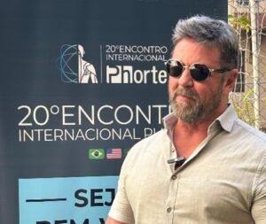 Professor Fábio Mazzonetto, homem de pele clara, cabelos grisalhos e barba espessa, usa óculos escuros e camisa de linho bege de mangas curtas. Ele está em pé em frente a um banner do “20º Encontro Internacional Phorte”, com os dizeres “Seja bem-vindo” e imagens em preto e branco de diversos palestrantes. O professor aparece gesticulando com as mãos, com microfone preso na camisa, em um ambiente externo cercado por folhagens e uma grade metálica ao fundo. O cenário remete ao espaço de recepção do evento, organizado pela Faculdade Phorte em São Paulo.