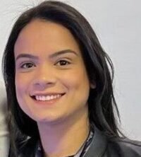 Luciana Capeloto, mulher jovem de pele clara e cabelos longos e lisos, pretos, aparece sorrindo suavemente para a câmera. Ela veste uma blusa escura com uma jaqueta preta por cima e usa um crachá com fita azul pendurado no pescoço, indicando participação no 20º Encontro Internacional Phorte. Ao fundo, a parede é branca e lisa, com iluminação clara. Alguns fios do cabelo estão levemente arrepiados no topo da cabeça, em uma pose espontânea e simpática.