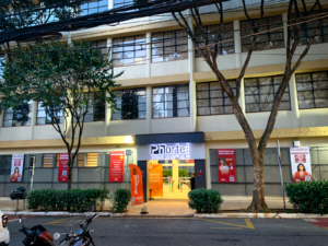 A fachada de um prédio amplo, com muitas janelas e paredes pintadas na cor bege. o centro da imagem está a entrada da Faculdade Phorte com a logo em cima da porta, iluminada.