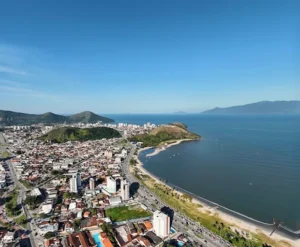 Vista aérea da cidade de Caraguatatuba, a imagem está dividida ao meio: de um lado podemos ver o mar e a orla da praia e, do outro, a parte urbana com prédios, casas, construções e pontuais áreas verdes
